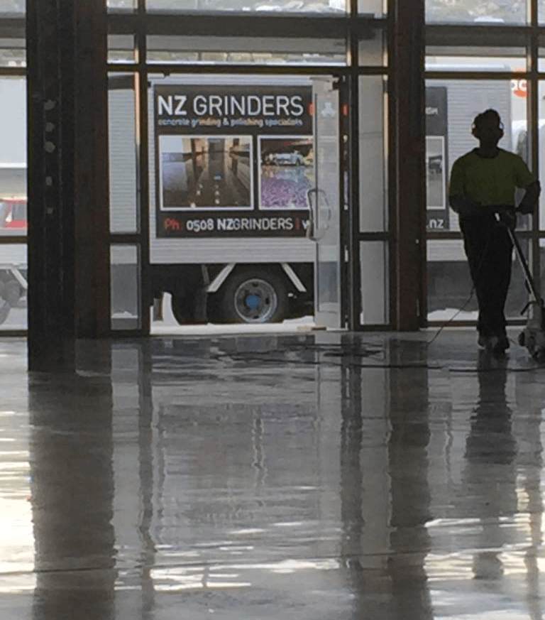 commercial polished floors - tauranga - nz grinders - warehouse polished concrete floor in a warehouse with a truck in the background shown through glass doors and a worker inside polishing the floor